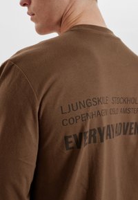 Camicia a maniche lunghe in cotone marrone con testo nero sul retro che presenta nomi di città e la scritta "EVERY ADVENTURE" in evidenza.