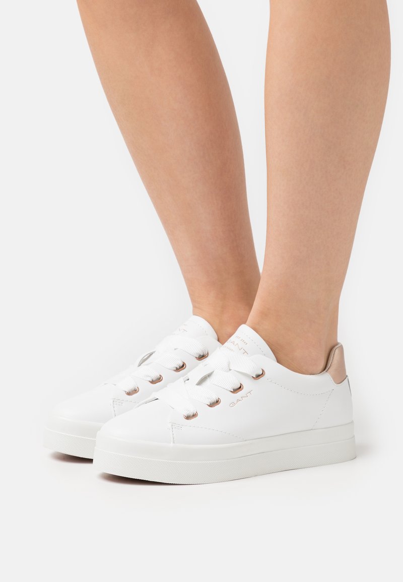 White sneakers featuring a smooth leather upper, round toe, and flat rubber sole. Accented with beige heel and metallic eyelets for laces.