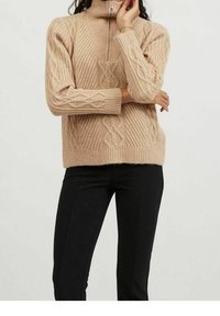 Maglione beige lavorato a maglia con colletto alto e dettagli con mezza zip, caratterizzato da motivi a treccia e texture a coste. Indossato con pantaloni neri aderenti.