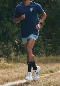 Camisa atlética azul marino, pantalones cortos azul claro, calcetines negros hasta la rodilla, zapatillas de correr blancas con acentos coloridos; al aire libre en un sendero.