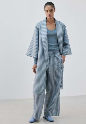 Kobieta w luźnej, niebiesko-białej, w paski kurtce w stylu kimono i spodniach, z niebieskim topem z długim rękawem oraz niebieskich butach wsuwanych, stoi na jednolitym tle.