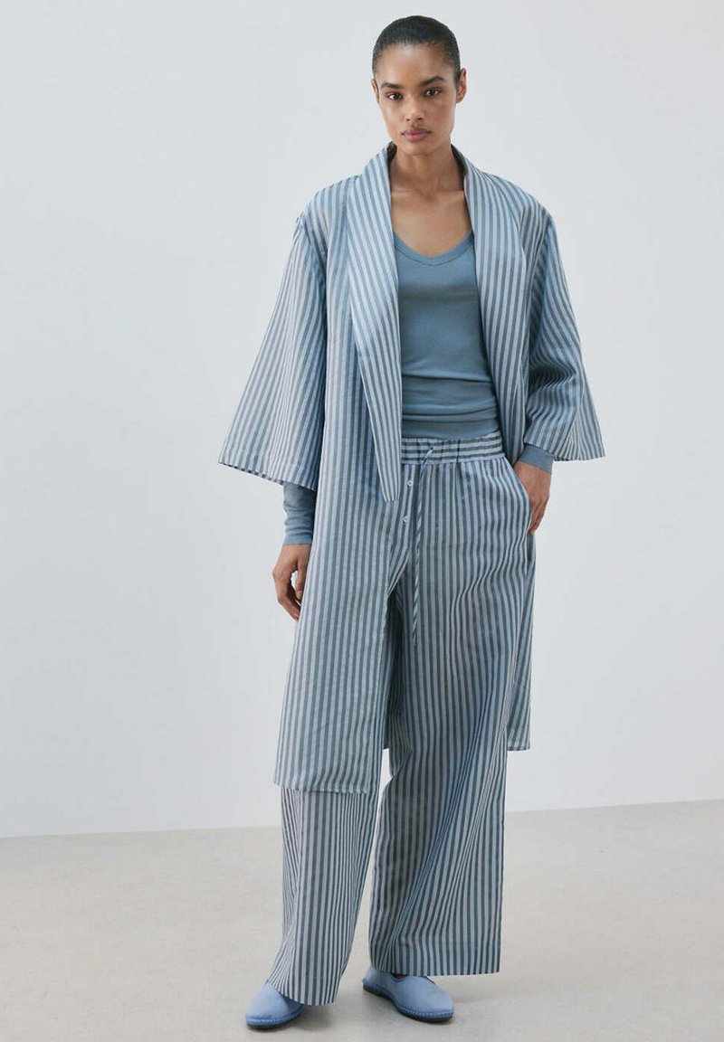 Femme portant une veste et un pantalon ample style kimono à rayures bleues et blanches, avec un haut bleu à manches longues et des chaussures bleu clair sans lacets, debout sur un fond uni.