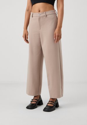 Pantaloni beige a gamba larga con una texture liscia, dotati di chiusura frontale con bottone, passanti per la cintura, abbinati a scarpe nere con tacco.