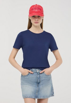 T-shirt a maniche corte blu navy abbinato a una minigonna di jeans azzurro. Il cappellino rosso presenta la scritta "NEWYORK" in lettere bianche nella parte frontale.