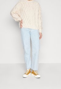 Kermainen, teksturoitu neule, jossa on sahalaitakuviota, yhdistettynä vaaleansinisiin suoriin farkkuihin ja keltaisiin korkeaseinäisiin tennoihin, joissa on valkoiset pohjat.