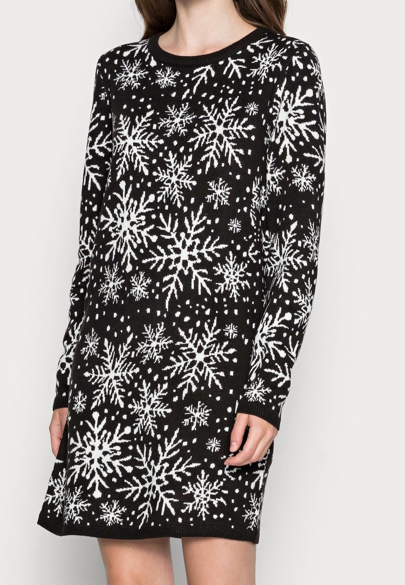 Femme portant une robe en tricot noire à manches longues avec des motifs de flocons de neige blancs et de petits points sur tout le tissu.