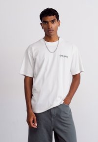 Tricou alb cu maneci scurte, cu guler rotund și logo-ul negru "DICKIES". Prezentând o croială lejeră și un material din bumbac neted. Poartă pantaloni șort gri.