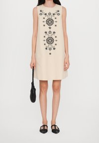 Vestido sin mangas beige con patrones florales bordados en negro, acompañado de un bolso tejido negro y zapatos planos negros. Silueta A-line simple.