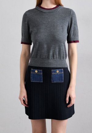 Femme portant un pull gris à manches courtes avec des bordures bordeaux et une jupe noire côtelée avec deux poches en denim bleu ornées de boutons dorés.