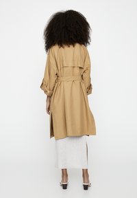 Lichtbruine trenchcoat met een strikceintuur, lange mouwen, een rugventilatie en een gladde textuur, gedragen over een witte geribde jurk.