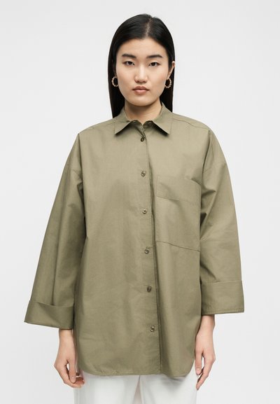 Γυναίκα με μακριά μαύρα μαλλιά που φορά χρυσούς κρίκους σκουλαρίκια και μια φαρδιά πουκαμίσα σε ελιά πράσινο χρώμα με τσέπη στο στήθος, στέκεται μπροστά σε απλό φόντο.