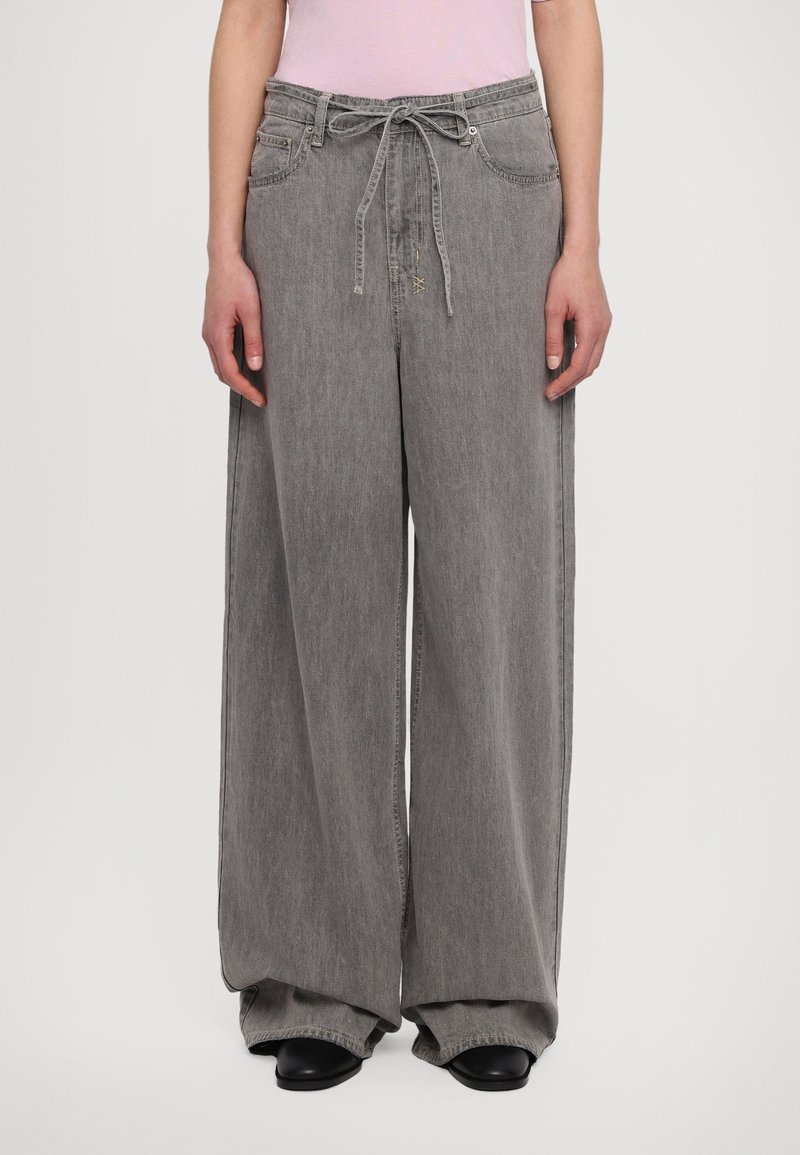 Personne portant un pantalon en denim gris ample avec une taille à cordon de serrage et des chaussures noires, montrée de la taille aux pieds sur un fond uni.
