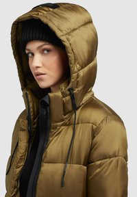 Veste rembourrée olive avec un col haut et une capuche réglable. Elle est dotée d'une fermeture éclair à l'avant et de cordons noirs, créant un aspect texturé.