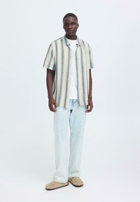 Korte mouwen, overhemd met knopen en horizontale strepen in pasteltinten, gedragen over een wit T-shirt, gecombineerd met lichtblauwe spijkerbroek en beige schoenen.