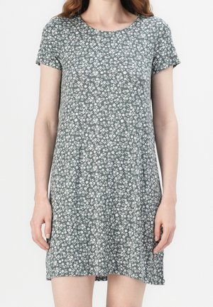 Femme portant une robe grise à manches courtes, longueur genoux, avec un motif floral blanc, debout devant un fond blanc uni.