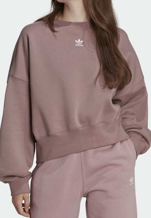 Žena má na sobě mauve mikinu Adidas a odpovídající tepláky, stojí s jednou rukou v kapse, viditelné logo Adidas na hrudi a stehně.