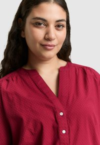 Bordeauxrote Bluse mit V-Ausschnitt, kurzen Ärmeln, Knopfleiste, strukturiertem Stoff mit dezentem Muster und gerafften Details an den Schultern.