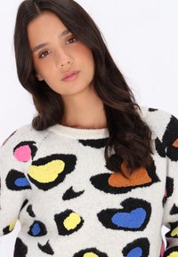 Pull blanc avec des motifs en forme de cœur noirs, bleus, jaunes, roses et orange ; texture douce et col rond ; coupe décontractée et ample.