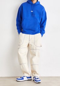 Felpa blu con cappuccio e tasca frontale, pantaloni a gamba larga crema con motivi di cucitura in contrasto e sneakers blu e bianche.