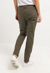 Persona caminando con pantalones verdes oliva, zapatillas blancas y una camiseta blanca contra un fondo blanco plano.