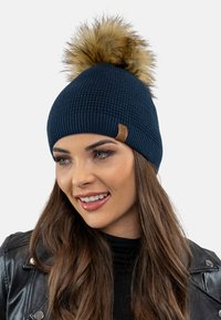 Căciulă tricotată de culoare bleumarin, cu un model texturat și un pom-pom din blană sintetică în vârf. Are un patch din piele cu brandul pe lateral.