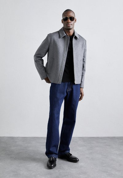Veste en mélange de laine grise avec un col chemise, fermeture éclair contrastante, t-shirt noir et Jean en denim bleu. Modèle portant des lunettes de soleil sombres et des chaussures.
