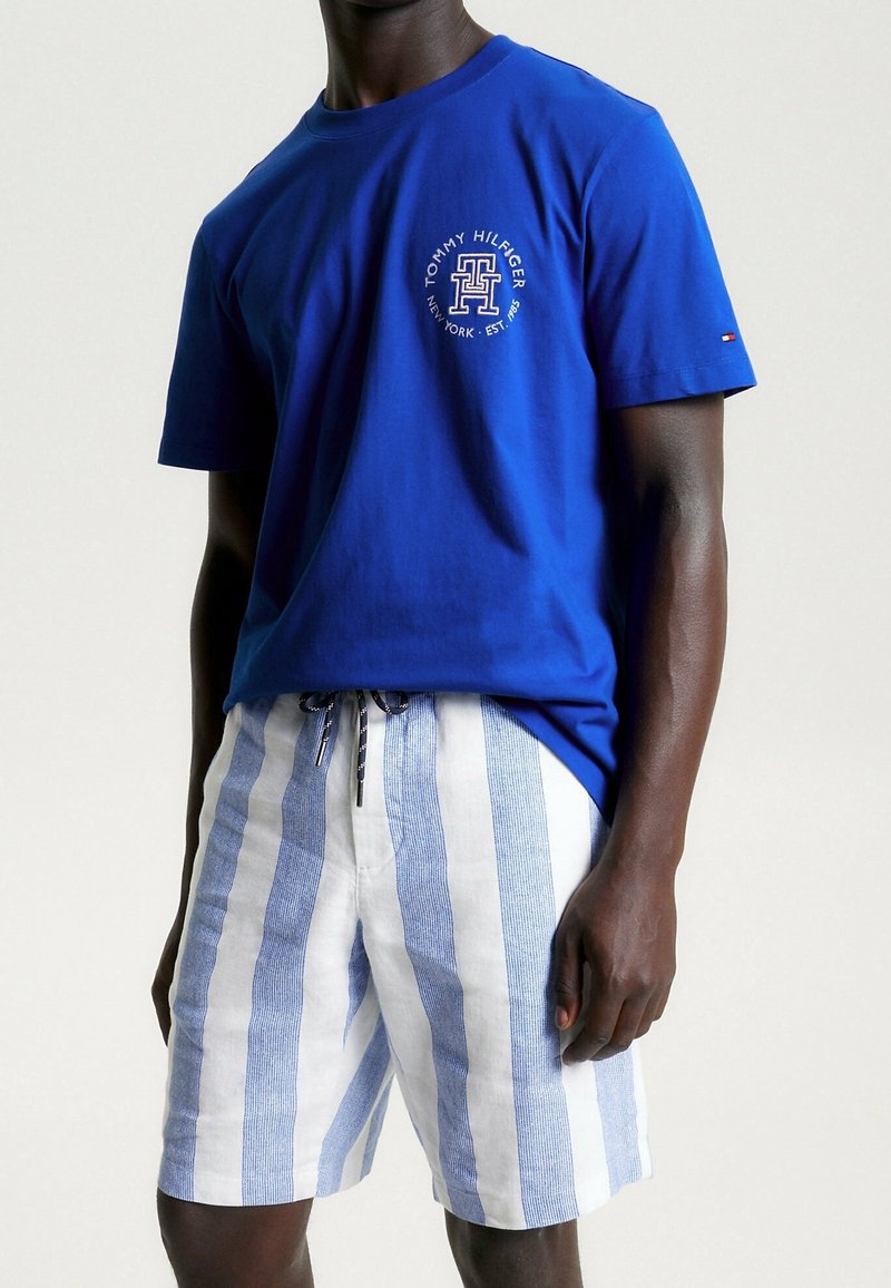 Camiseta de algodón azul con cuello redondo y estampado de logo; combinada con pantalones cortos de lino rayados en blanco y azul claro con cintura ajustable.