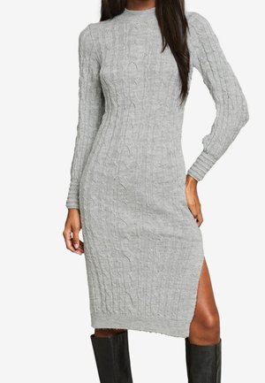 Robe midi gris ajustée en maille torsadée à manches longues, col montant et fente latérale, portée avec des bottes noires montantes au genou.