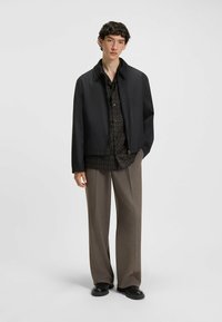 Black bomber jacket over a dark patterned shirt, paired with light brown wide-leg trousers and black shoes. Smooth fabrics, relaxed fit.