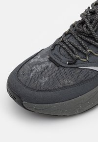 Zapatilla deportiva gris con parte superior de malla texturizada, puntera reforzada y superposiciones con patrones. La suela es gruesa y tiene un diseño estriado.