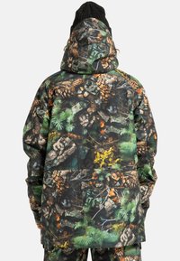 Osoba w kapturze, ubrana w kurtkę i spodnie w szczegółowy leśny wzór kamuflażowy z motywem szyszek, gałęzi i zielonego liścia, widziana z tyłu.