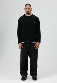 Zwarte trui met textuur en lange mouwen, wit ondershirt, losse zwarte broek en stevige zwarte schoenen met gekleurde veters. Subtiele logodetails.