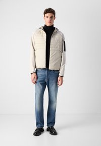 Light beige puffer jacket with a hood, featuring a textured upper, black accents on the sleeves, paired with blue jeans and black shoes.