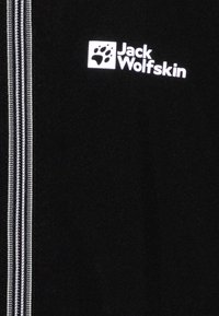 Černá tkanina s bílým vertikálním zipem na levé straně a bílým logem Jack Wolfskin s otiskem tlapky v pravém horním rohu.