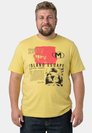 Bärtiger Mann trägt ein gelbes T-Shirt mit dem Schriftzug „Island Escape“ und Palmen-Grafiken, steht vor einem schlichten hellen Hintergrund.