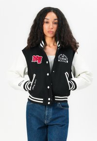 Giacca varsity bicolore in bianco e nero con logo ricamato "NY", maniche bianche, polsini a righe e testo "Tommy Jeans" lungo l'orlo.