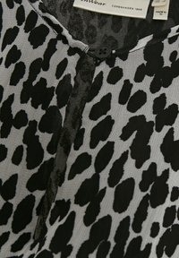 Tissu imprimé léopard noir et gris avec un col rond, une fermeture à un bouton noir unique, et une étiquette de vêtement visible.