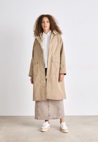 Beige waterdichte trenchcoat met een trekkoordcapuchon, gouden knopen, twee voorzakken en een A-lijn silhouet. Gemaakt over een licht shirt.