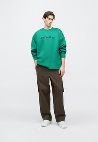 Πράσινο φούτερ με το κείμενο "JUST DO IT." σε μαύρο χρώμα, συνδυασμένο με καφέ παντελόνι cargo. Η εμφάνιση ολοκληρώνεται με λευκά αθλητικά παπούτσια.