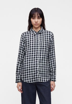 Femme aux cheveux noirs et lisses porte une chemise à carreaux noire et blanche boutonnée et un pantalon sombre, debout devant un fond blanc uni.