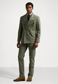 Šviesiai alyvuogių žalia dvieilė dvieilė kostiumas su įpjautomis atlapinėmis, dviem priekinėmis kišenėmis, derančiomis kelnėmis ir rudos spalvos laisvalaikio batais. Po apačia dėvima languota marškinėliai.