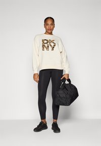 Krém színű pulóver leopárdmintás logóval, fekete leggings, fekete sportcipő és fekete duffel táska. Simább textúrák és laza dizájn.