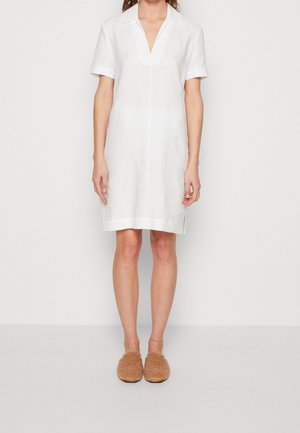 Woman wearing a short-sleeve, knee-length white dress with a collar and tan fuzzy slip-on shoes, standing against a plain background.