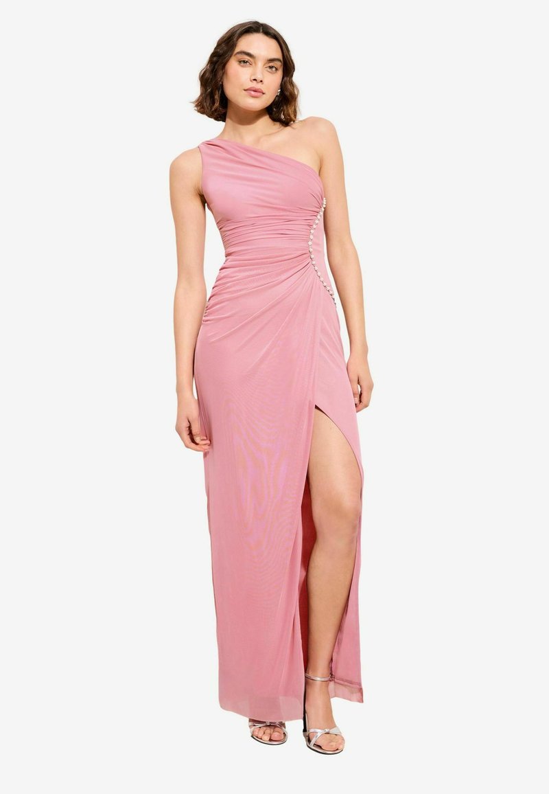 Kvinde iført en pink, asymmetrisk aftenkjole med draperinger, sideslids og perleknapdetalje, kombineret med sølvsandaler med stropper.