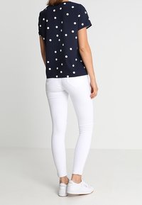 Marineblauwe blouse met witte polka dots, korte mouwen; gecombineerd met witte spijkerbroek en witte sneakers. Soepele textuur, casual pasvorm.