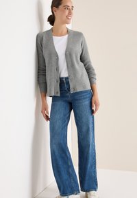 Hellgraue, gerippte Strickjacke mit Knöpfen, getragen über einem weißen T-Shirt und kombiniert mit weit geschnittenen blauen Jeans, vor einem neutralen Hintergrund.