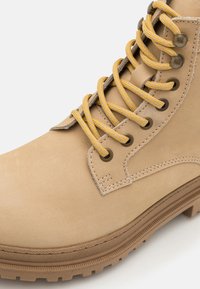 Bottine beige en cuir lisse, avec des lacets à motifs jaunes et gris, œillets en métal et semelle en caoutchouc texturé.