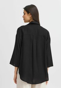 Camicia nera, leggera, con vestibilità ampia. Presenta maniche lunghe, colletto e spallina sul retro con una leggera piega per un dettaglio di design aggiuntivo.