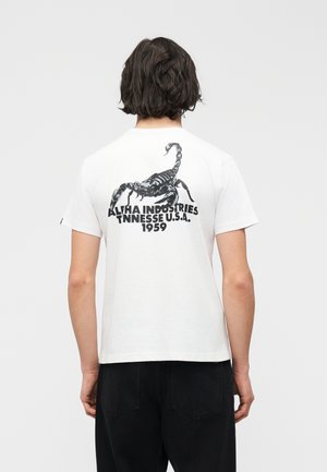 Personne aux cheveux foncés portant un t-shirt blanc avec un grand motif de scorpion noir et le texte "Alpha Industries Tennessee U.S.A. 1959" au dos.