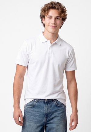 Junger Mann mit lockigem Haar, der lächelt, trägt ein weißes kurzärmeliges Poloshirt und blaue Jeans und steht vor einem schlichten hellen Hintergrund.